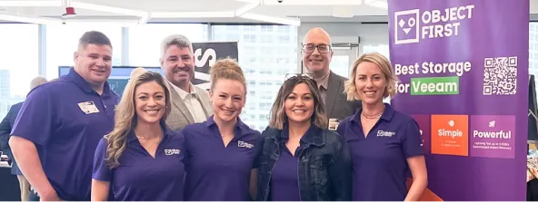 A group of seven people posing together in a brightly lit office or conference setting, with four women wearing matching purple shirts, standing in front of a purple banner that reads "OBJECT FIRST" and promotes storage solutions for Veeam.