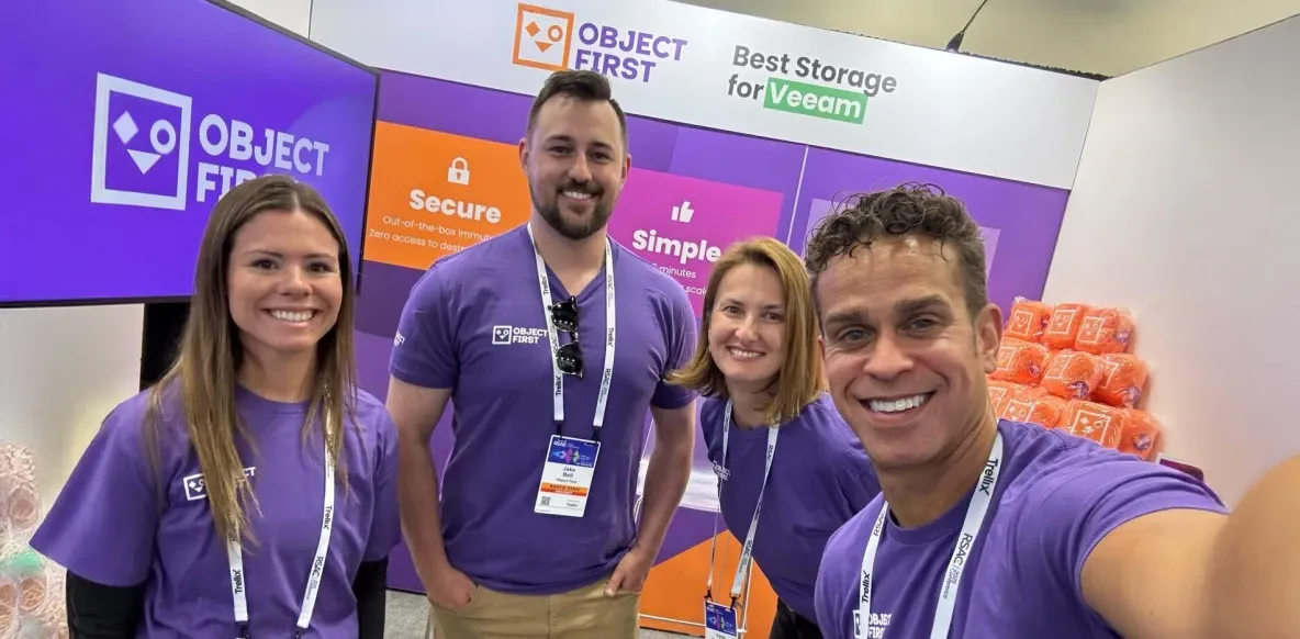 Um grupo de sete pessoas posando juntas em um ambiente de escritório ou conferência bem iluminado, com quatro mulheres usando camisas roxas combinando, em frente a um banner roxo com o texto 'OBJECT FIRST', promovendo soluções de armazenamento para Veeam.