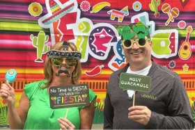 Deux personnes habillées de manière festive avec des accessoires colorés, portant des lunettes amusantes et posant devant un décor vibrant à thème de fête.