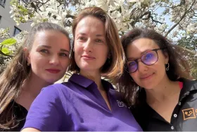 Tre donne all'aperto sotto un albero in fiore, sorridendo alla fotocamera, con una che indossa occhiali da sole viola.