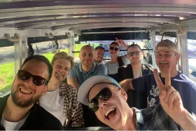 Group of smiling people riding together in an open vehicle, making cheerful gestures and posing for a photo.