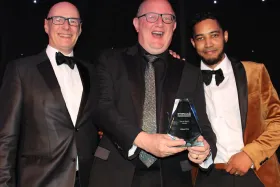 Three men dressed formally in tuxedos, two of them holding an award and celebrating on stage.