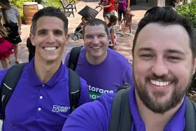 Three men taking a selfie outdoors, all smiling, wearing purple shirts, and carrying backpacks.