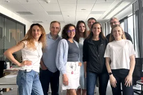 Un groupe de sept personnes posant ensemble dans un environnement de bureau, debout de manière décontractée et souriant.