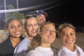 A group of five people smiling at the camera in an outdoor setting at night, with bright lights in the background.